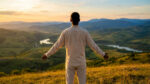 Back view of a person in light clothes on a golden hour hilltop, arms open to a serene landscape with hills, mountains, and river.