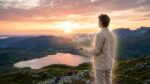 Un homme en contemplation face à un lever de soleil sur des montagnes et un lac. Des étincelles lumineuses émanent de ses mains ouvertes.