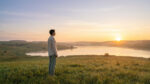 Un homme debout dans une prairie au lever du soleil, observant un lac paisible dans une atmosphère sereine.